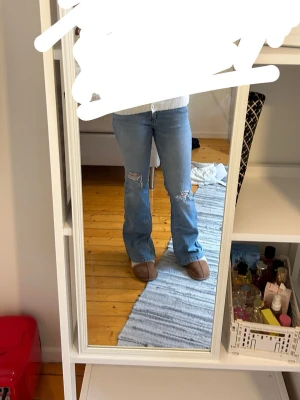 Ljusblå lågmidjade bootcut jeans med slitningar - Säljer ett par ljusblå lågmidjade bootcut jeans med slitningar på knäna. Jeansen har klassisk femficksdesign och är tillverkade i ett mjukt denimtyg. Perfekta för dig som gillar en avslappnad och trendig stil.