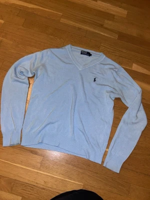 Ljusblå v-ringad tröja Polo Ralph Lauren - Ljusblå v-ringad tröja från Polo Ralph Lauren med klassisk broderad logga på bröstet. Tröjan har långa ärmar och ribbade muddar vid ärmslut och nederkant. Perfekt för en stilren och avslappnad look. Fint skick, lite noppig.