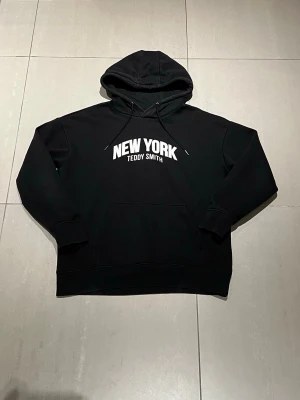Svart hoodie Teddy Smith New York - Svart hoodie från Teddy Smith med stor vit text 'NEW YORK' på bröstet. Klassisk känguruficka framtill och justerbar huva med snören. Tillverkad i mjukt bomullsmaterial som är skönt att bära. Perfekt för en chill och urban look.