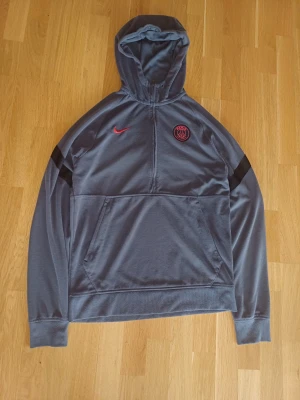 Grå PSG hoodie från Nike - Snygg PSG quarter zip-hoodie från Nike i storlek S. Har mest legat i garderoben och inte använts på ett tag.  Plagget är i fint skick överlag finns en liten blå fläck och Nike broderiet på loggan är lite snett, men inget som märks när man har den på.  Väldigt bekväm och lätt att styla