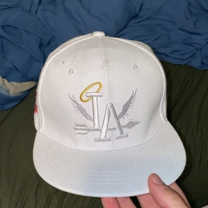 Vit LA snapback med broderi - Snygg vit snapback-keps med broderat LA-logo framtill, dekorerad med vingar och en gul gloria. På sidan finns färgglatt broderi med texten 'World Champs' och palmer. Justerbar passform med plastspänne. Perfekt för dig som gillar streetwear och sportig stil.