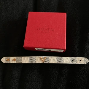 Valentino Garavani armband i skinn - Snyggt armband från Valentino Garavani i vitt och svart rutigt mönster på ovansidan och orange på undersidan. Armbandet är i skinn och har ett guldigt V i mitten samt tryckknappar i guld. Kommer med original röd ask. lådan ingår inte