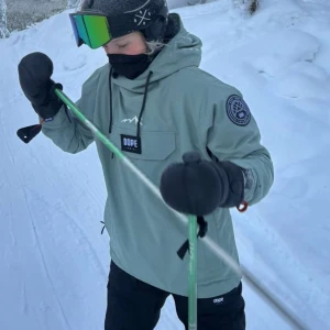 Snowboard jacka Dope  - Snygg ljusgrön vindjacka från Dope med stor magficka, huva med snörning och patch på ärmen. Jackan har en clean look med svart logotyp framtill och justerbara muddar. Perfekt för dig som gillar att åka skidor eller snowboard. Använd en veckas resa, inga slitage. Nypris 2099