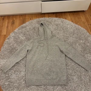 Grå stickad hoodie med huva - Mysig grå stickad hoodie med huva och dragsko. Tröjan har en klassisk känguruficka framtill, ribbade muddar vid ärmslut och nederkant samt lång ärm. Perfekt för kyliga dagar när du vill vara både bekväm och stilren.