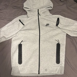 Grå Nike Tech Fleece hoodie M - Nike Tech Fleece hoodie i ljusgrått med svarta detaljer och dragkedjor. Tröjan har hel dragkedja, två stora fickor med svarta dragkedjor och en mindre ficka på ärmen. Klassisk Nike-logga på bröstet och skön huva. Perfekt för chill eller träning.Dock har den en fläck vid midjan så man vet