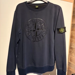 Blå Stone Island sweatshirt - Säljer en mörkblå sweatshirt från Stone Island med stort logomärke framtill och klassisk patch på ärmen. Tröjan har rund halsringning och långa ärmar, perfekt för dig som gillar streetwear och snygga detaljer.