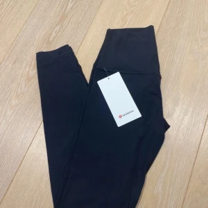 Svarta ribbade leggings från Lululemon - Svarta ribbade leggings från Lululemon, modell Align Ribbed HR Pant 28”. Hög midja och smörmjuk känsla med fyrvägsstretch. Tillverkade i svettavvisande Nulu-tyg med inslag av Lycra för extra stretch. Dold ficka i linningen för nyckel eller kort.