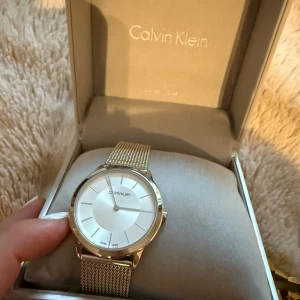 Silverfärgad Calvin Klein klocka - Stilren klocka från Calvin Klein med rund urtavla i vitt och silver. Klockan har ett mesharmband i metall och minimalistisk design med tunna markeringar. Perfekt accessoar för dig som gillar en clean och modern look.