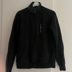 Svart fleecejacka Sail Racing - Svart fleecejacka från Sail Racing med hög krage och hel dragkedja framtill. Jackan har en bröstficka med dragkedja och logga, samt två sidofickor. Perfekt för kyliga dagar och enkel att matcha med allt.