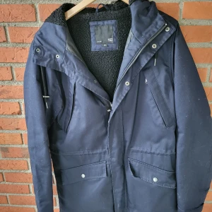 Mörkblå parkasjacka med huva, 157 Storlek M passar L - Mörkblå parkasjacka från 157 med huva och mysigt svart teddyfoder. Jackan har dragkedja och tryckknappar framtill, stora fickor med lock och innerficka. Perfekt för kyliga dagar med sin långa modell och värmande material.