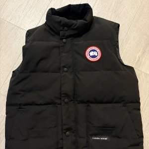 Canada Goose Väst - Svart dunväst från Canada Goose med hög krage, två stora fickor framtill och klassisk logga på bröstet. Västen har både dragkedja och knappar framtill för extra värme och stil. Perfekt för lager-på-lager under kyliga dagar. Canada Goose Väst i mycket bra skick, inga skador, inga fläckor, som ny. Kvitto finns fortfarande kvar.