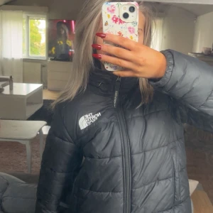 Svart pufferjacka från The North Face - Säljer en svart pufferjacka från The North Face i storlek M. Jackan har hög krage, dragkedja framtill och logga på bröstet. Finns en luva gömd i kragen som man öppnar med dragkedjan på kragen. Säljer då den inte kommer till användning längre.