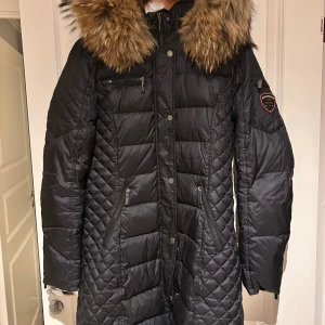 Svart lång dunjacka Rockandelle - Snygg svart lång dunjacka från Rockandelle med stor fluffig fuskpäls runt kragen. Jackan har quiltad design, flera dragkedjefickor och tryckknappar framtill. Märkespatch på ärmen och figurnära passform. Perfekt för kalla vinterdagar.