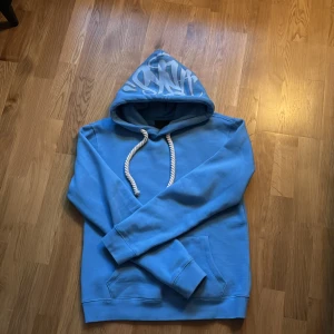 Blå Hoodie från syna world  - Ljusblå hoodie från Syna World. I fint skick. Har grova, flätade snören och graffiti-text på huvan. Följer med en Syna World-påse, en nyckelring och klistermärken. Såklart kan pris ändras, nypris 1200kr
