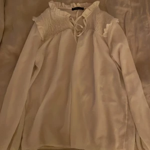 Vit blus med volang från Trendyol - Supersöt vit blus från Trendyol med volangdetaljer vid halsen och axlarna, samt smockade partier upptill och vid ärmsluten. Blusen har långärmade ärmar och knytning vid halsen. Perfekt för dig som gillar romantisk och feminin stil.