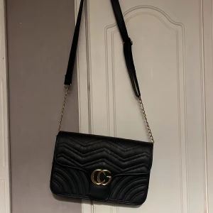 Svart axelväska med gulddetaljer - Snygg svart axelväska med quiltad design och guldfärgat CG-spänne framtill. Väskan har kedjerem och justerbar axelrem i skinnimitation. Perfekt för dig som vill ha en trendig och stilren accessoar.