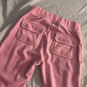Rosa mjukisbyxor Juicy Couture XXS - Säljer ett par ikoniska rosa mjukisbyxor från Juicy Couture i storlek XXS. Byxorna har bred resår i midjan, snörning framtill och stora fickor med lock och silverfärgade knappar bak. Mjukt material och klassisk Juicy-stil.