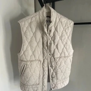 Beige quiltad väst från Zara XS - Quiltad beige väst från Zara i storlek XS. Västen har hög krage, tryckknappar framtill och två stora fickor. Den har ett snyggt mönster med sydda rutor och är tillverkad i ett mjukt bomullsmaterial. Perfekt att styla över en hoodie eller långärmad tröja.