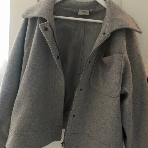 Grå kappa från ZINK - Snygg grå kappa från ZINK med klassisk krage och stor bröstficka. Jackan har dragkedja och tryckknappar framtill, perfekt för lager-på-lager. Materialet är mjukt och känns som ull, vilket gör den både varm och stilren. 