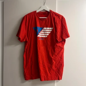 Röd Puma t-shirt med tryck XL - Röd t-shirt från Puma i bomull med klassiskt crewneck och stort grafiskt tryck i blått och vitt framtill. Skön passform och perfekt för dig som gillar sportig stil. Snygg att matcha med jeans eller shorts.