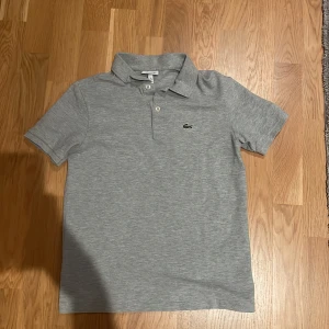  Grå pike från Lacoste - Snygg grå pikétröja från Lacoste med klassisk krage och två knappar framtill. Tröjan har korta ärmar och den ikoniska gröna krokodilloggan broderad på bröstet. Perfekt för dig som gillar stilrena och sportiga plagg. Vänligt länge sedan jag hade på mig den sist, använt max 3 gånger eftersom den redan var liten för mig när jag fick den som present. Storlek 14/164 fast kan passa 15/170 beronde på hur slim fit du vill ha den. KOM MED FRÅGOR OCH PRISFÖRSLAG ‼️