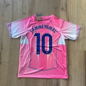 Barça Lamine Yamal rosa matchtröja - Säljer en officiell FC Barcelona fotbollströja från Nike med Lamine Yamal och nummer 10 på ryggen. Tröjan är rosa med vita och ljusblå detaljer, har korta ärmar och V-ringad krage. Materialet är lätt och andas, perfekt för träning eller match. Spotify-logga på bröstet.