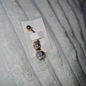 Navelpiercing - Snygg oanvänd navelpiercing i guldton med två stora, glittriga stenar som fångar ljuset. Stenarna är klara och sitter i en rund infattning, vilket ger ett lyxigt och trendigt intryck. Perfekt för dig som vill ha något som verkligen syns och glittrar.