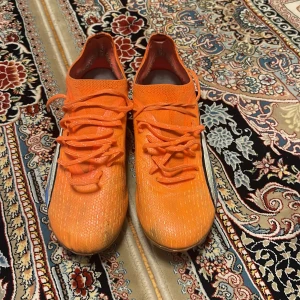 Puma ultra pro orange - Säljer ett par grymma orangea fotbollsskor från Puma med snörning och platt sula med dobbar. Skorna har en snygg vit och blå detalj på sidan och är tillverkade i syntetmaterial för bästa prestanda på planen. Perfekt för dig som vill sticka ut med färg och stil.