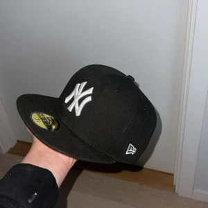Svart New Era Yankees keps 59FIFTY - Säljer en svart New Era 59FIFTY keps med New York Yankees-logga broderad i vitt framtill. Klassisk flat peak och broderad MLB-logga bak. Kepsen är i fast storlek och tillverkad i ett slitstarkt material.