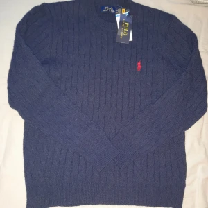 Mörkblå kabelstickad tröja Polo Ralph Lauren - Kabelstickad mörkblå tröja från Polo Ralph Lauren med rund halsringning och den klassiska röda logotypen broderad på bröstet. Tröjan har långa ärmar och ribbade muddar vid ärmslut och nederkant. Perfekt för en stilren och avslappnad look.