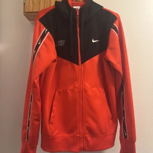 Nike röd och svart hoodie med dragkedja - Nike hoodie i rött och svart med hel dragkedja, huva och snörning. Snygga vita Nike-loggor på bröstet och längs ärmarna. Sportig look med svarta detaljer upptill och på huvan. Perfekt för dig som gillar streetwear och sportig stil.
