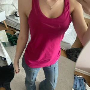 Rosa Dri-Fit linnetopp från Nike - Säljer ett sportigt rosa Dri-Fit linne från Nike med klassisk logga framtill. Linnet har racerback och är tillverkat i ett funktionsmaterial som andas, perfekt för träning. Slim fit-modell som sitter snyggt och bekvämt.