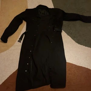 Svart lång rock från Pieces - Lång svart rock från Pieces med klassisk krage och knäppning framtill. Jackan har ett bälte i midjan och raka ärmar. Perfekt för dig som gillar en stilren och tidlös look. Passar dig som vill ha en snygg och enkel jacka till höst och vinter.