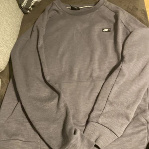 Grå sweatshirt från Nike i storlek M - En stilren grå sweatshirt från Nike med klassisk rund hals och diskret Nike-logga på bröstet. Tröjan har långa ärmar och ribbade muddar vid ärmslut och nederkant. Perfekt för en avslappnad och sportig look.