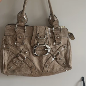 Beige handväska från Guess med detaljer - Snygg beige handväska från Guess med GG-logga över hela väskan, silverfärgade metalldetaljer och spännen. Väskan har flera remmar och dekorativa nitar samt präglade partier som ger en cool look. Perfekt för dig som gillar statement-accessoarer.
