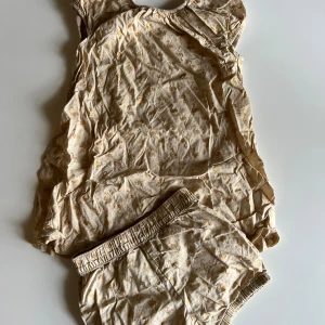 Beige tröja och shorts med guldmönster från H&M - Säljer ett par söta beiga shorts och tröja från H&M i storlek 92. Shortsen har ett glittrigt guldmönster med flamingos och resår i midjan för skön passform. Perfekta för varma dagar och riktigt gulliga till sommaren.