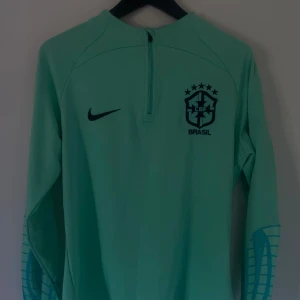 Brasil långärmad träningströja Nike - {FAST PRIS} Snygg ljusgrön långärmad fotbollströja från Nike med Brasil-logga och svart swoosh på bröstet. Tröjan har halv dragkedja framtill och coolt mönster på ärmarna. Perfekt för dig som gillar landslagsstil och sportig look.