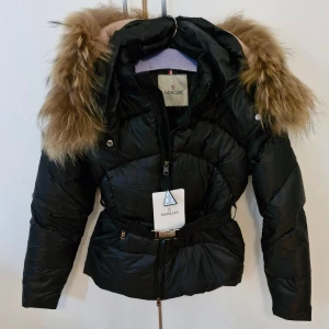 Svart Moncler dunjacka med päls Stl S=1 - Svart dunjacka från Moncler med fluffig päls på huvan, snyggt bälte i midjan och dragkedja framtill. Jackan har quiltad design, två fickor med dragkedja och en cool silvrig spänne. Perfekt för kalla dagar och riktigt stilren look. Storlek 1 = Small. Säljes p.g.a viktuppgång😒 fått den i present för 1 månad sedan så går ej att lämna tillbaka mm.