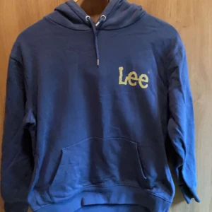 Blå hoodie från Lee med logga - Säljer en blå hoodie från Lee med broderad gul logga på bröstet. Tröjan har huva med snörning och en stor magficka. Perfekt för dig som gillar en avslappnad och stilren look.