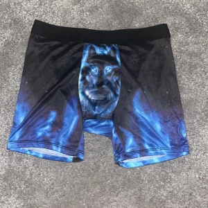 Svarta och blå shorts med vargmotiv - Coola shorts i svart och blått med ett stort vargmotiv framtill. De har en elastisk midja och är tillverkade i ett stretchigt material av polyester och spandex. Perfekta för dig som vill sticka ut med ett unikt tryck och bekväm passform.
