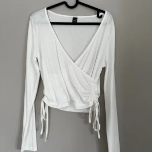 Vit omlottopp med snörning från Shein - Säljer en vit omlottopp från Shein med ribbad struktur och långa ärmar. Toppen har v-ringning och snörning i sidan. Aldrig använd 