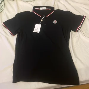 Svart Moncler piké med röd/vit detalj - Svart pikétröja från Moncler i storlek M. Helt ny med prislapp. Pris kan diskuteras vid snabb affär.