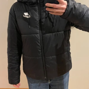 Svart pufferjacka från Nike - Säljer en svart pufferjacka från Nike med huva och dragsko. Jackan har en enkel look med diskret Nike-logga på bröstet, dragkedja framtill. Bruksanvänd, inga defekter. Storlek M. 
