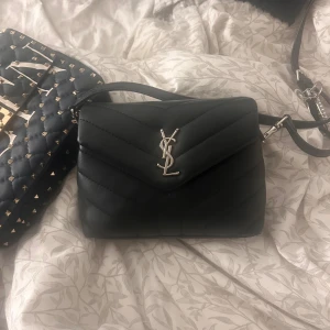 Svart quiltad axelväska från YSL - Säljer en svart quiltad axelväska från Saint Laurent med ikoniskt YSL-emblem i silver framtill. Väskan är tillverkad i skinn och har justerbar axelrem samt innerfack med dragkedja. Snygg och stilren modell som lyfter varje outfit.