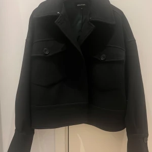 Svart jacka från Meotine - Stilren svart jacka från Meotine med stora bröstfickor och knappar framtill. Jackan har en boxig, croppad passform och klassisk krage. Perfekt för lager-på-lager och enkel att styla till olika outfits. Pälskragen finns också, bara inte med på bilden