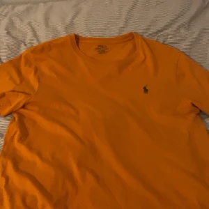 Orange t-shirt från Polo Ralph Lauren - Snygg orange t-shirt från Polo Ralph Lauren i modellen Custom Slim Fit. Klassisk kortärmad design med rund hals och ikoniska broderade loggan på bröstet. Tillverkad i mjuk bomull som känns skön mot huden. Perfekt för dig som gillar stilrena och färgstarka plagg.