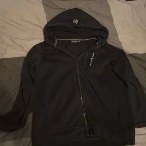 Svart hoodie från Sail Racing - Svart hoodie från Sail Racing med dragkedja framtill och diskret logga på bröstet. Tröjan har huva och långa ärmar, perfekt för chill dagar. Enkel och stilren design som funkar till det mesta.