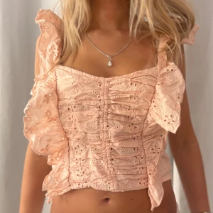 Ljusrosa blus med volanger och broderi - Supersöt ljusrosa blus med breda axelband och volanger längs sidorna. Toppen har broderade blommor och är croppad med rynkade detaljer framtill. Perfekt för dig som gillar romantisk och feminin stil. Materialet känns lätt och luftigt.insydd i midjan