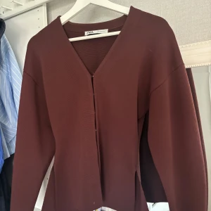 Brun v-ringad kofta från Zara - Snygg brun kofta från Zara med v-ringad hals och långärmad design. Koftan har en stilren look med slitsar i sidorna och är tillverkad i ett mjukt, strukturerat material som ger en skön känsla. Perfekt för dig som gillar minimalistisk stil.