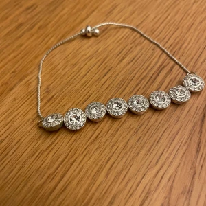 Silverfärgat armband med glittriga stenar - Snyggt armband i silverfärg med åtta runda länkar, varje länk har en stor klar sten i mitten och mindre stenar runt om som ger extra glittrig effekt. Justerbar kedja med små silverkulor. Perfekt för dig som gillar bling och vill sticka ut.